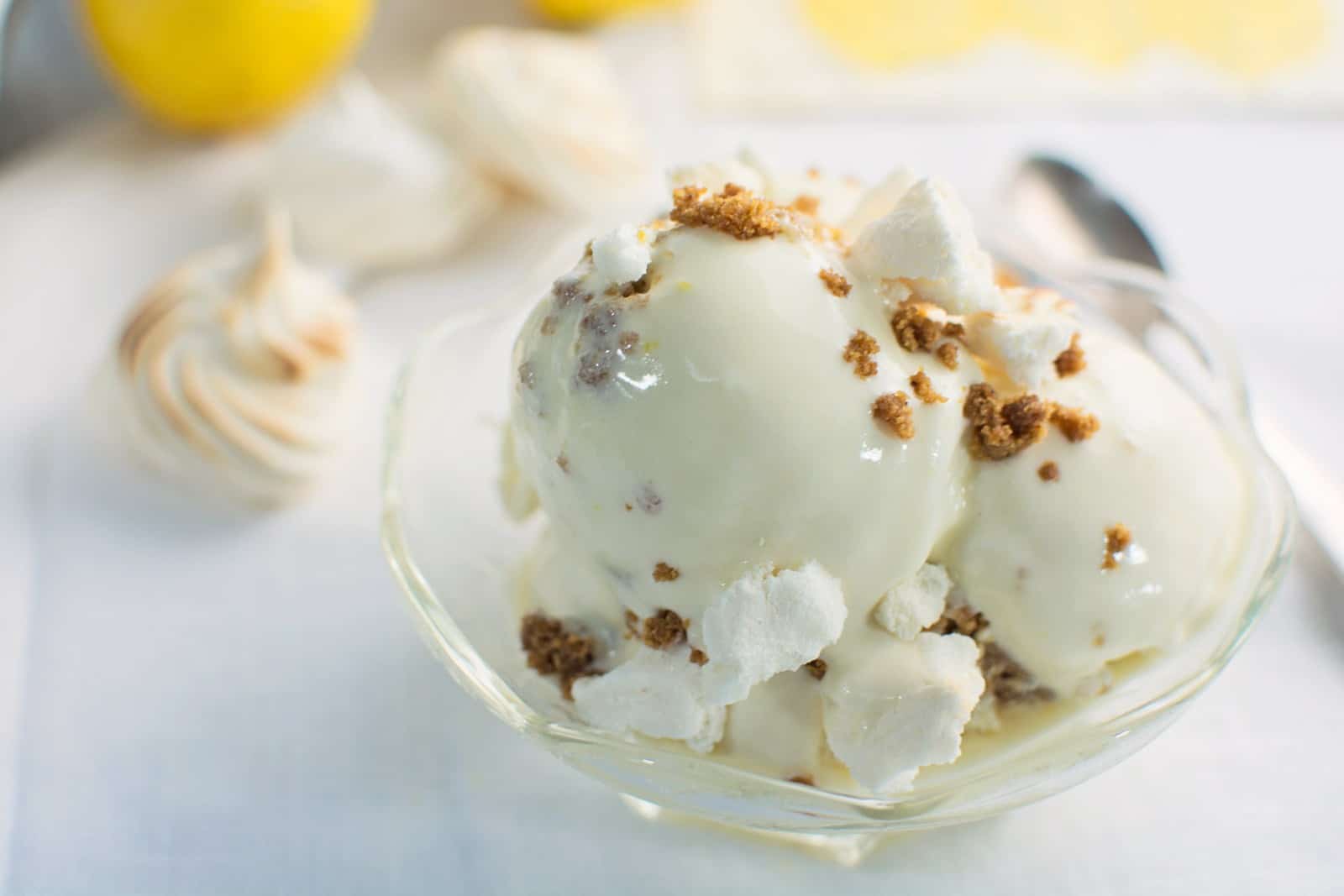 A glass bowl filled with scoops of creamy Lemon Pie Ice Cream topped with crumbled cookies and pieces of meringue, with a spoon and extra meringues in the background.