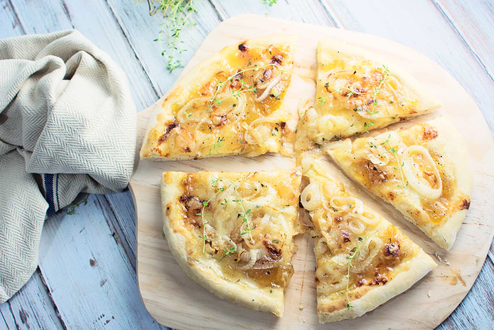 A sliced Onion Brie Pizza with herbs sits on a wooden board, next to a beige cloth napkin on a whitewashed wooden surface.
