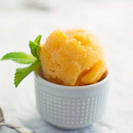 A scoop of apricot sorbet in a white textured cup, garnished with a sprig of fresh mint, set on a light-colored surface.