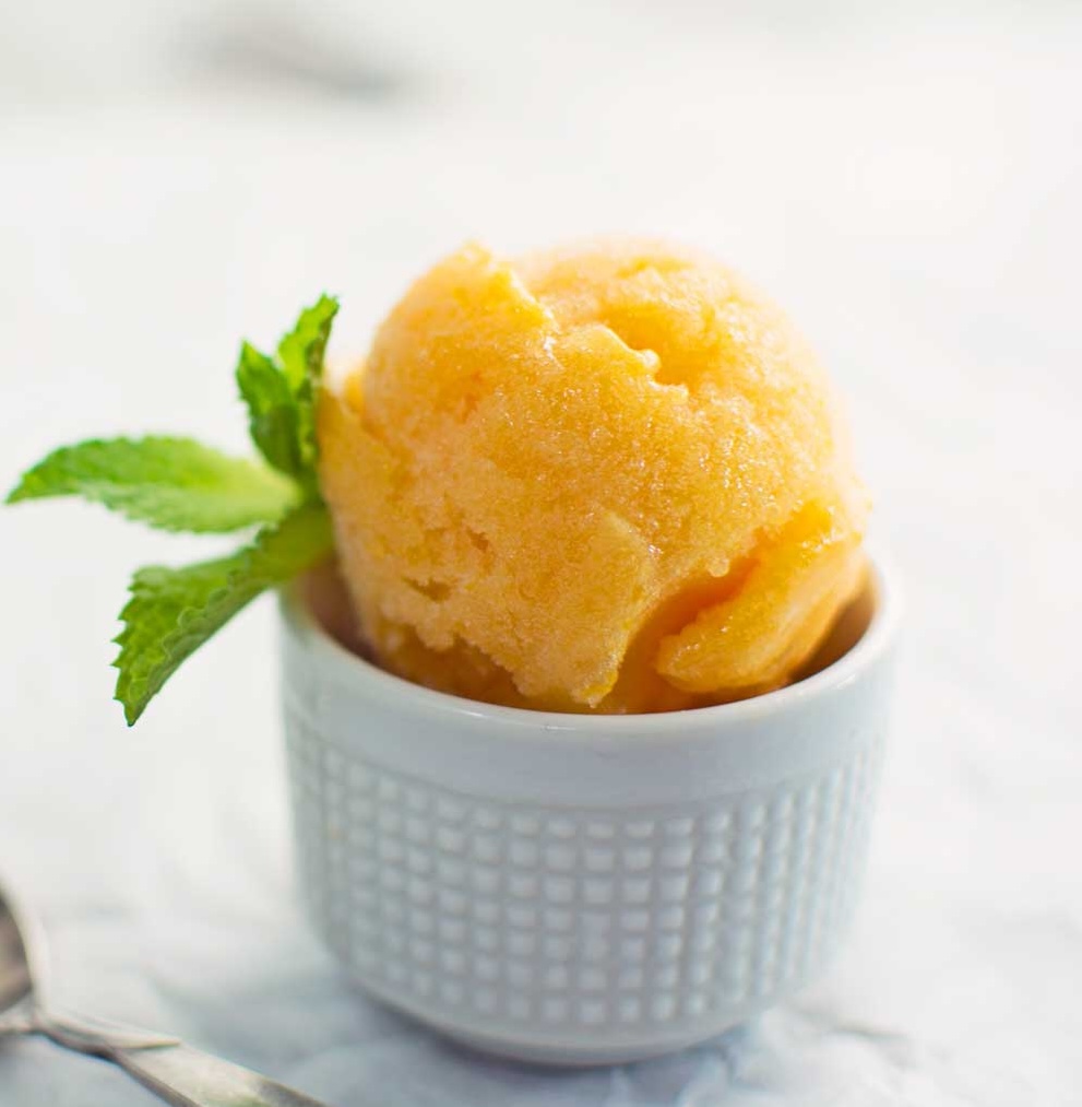 A scoop of apricot sorbet in a white textured cup, garnished with a sprig of fresh mint, set on a light-colored surface.