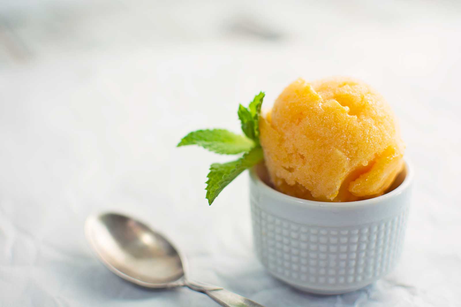A scoop of apricot sorbet in a small white cup, garnished with fresh mint leaves, sits next to a silver spoon on a light, textured surface.