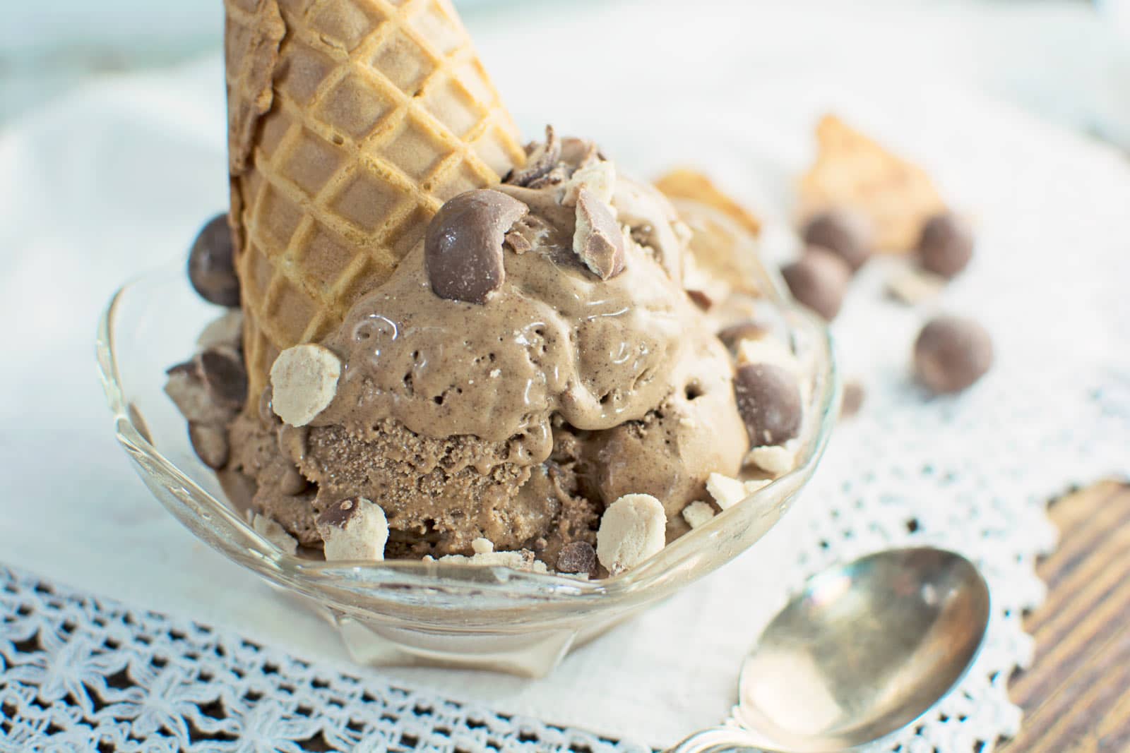 A glass bowl filled with malt ice cream, topped with chocolate chips and pieces of waffle cone, with a whole ice cream cone placed upside down. A spoon and lace napkin are beside the bowl.