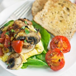 A plate with a fluffy omelette topped with mushrooms, cherry tomatoes, and herbs, served alongside fresh spinach, grilled tomato halves, and two slices of whole grain toast. A fork and a glass are partly visible in the background.