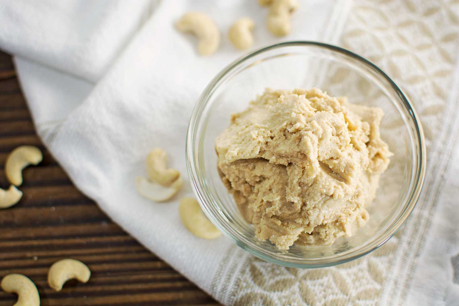 A glass bowl filled with creamy Cashew Butter sits on a patterned towel, surrounded by scattered whole cashews on a wooden surface.