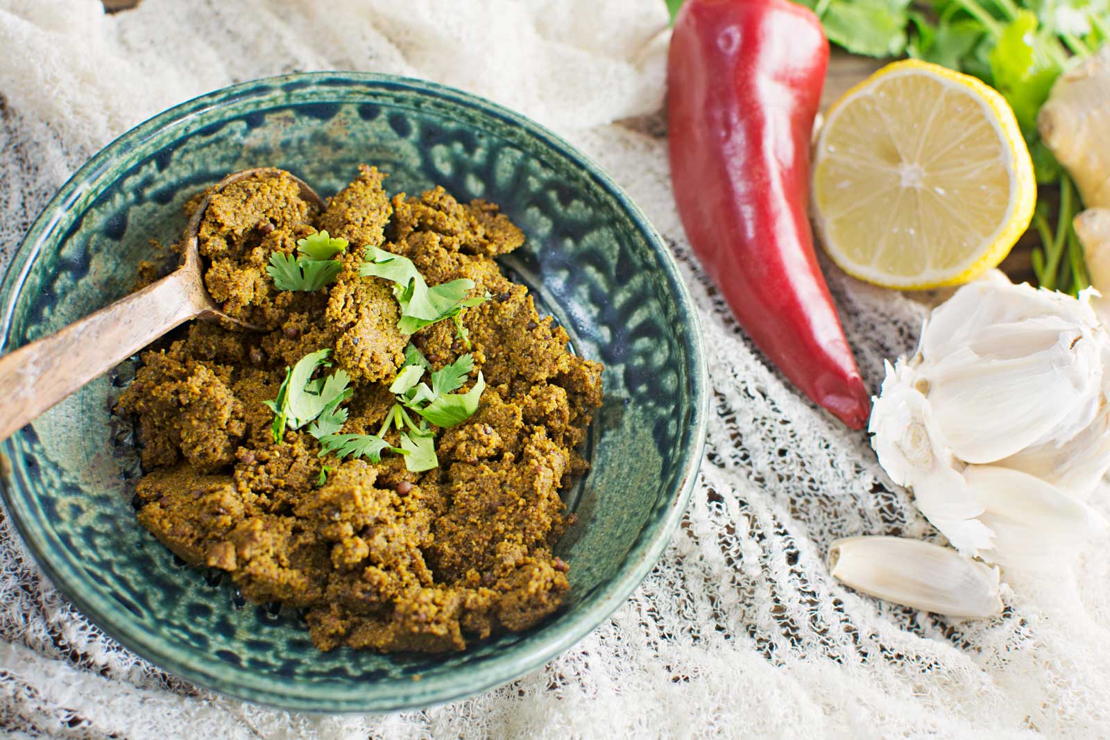 A green ceramic bowl filled with spicy brown curry paste garnished with cilantro beside a wooden spoon. Nearby are a red chili, half a lemon, garlic cloves, ginger, and fresh cilantro on a textured cloth.