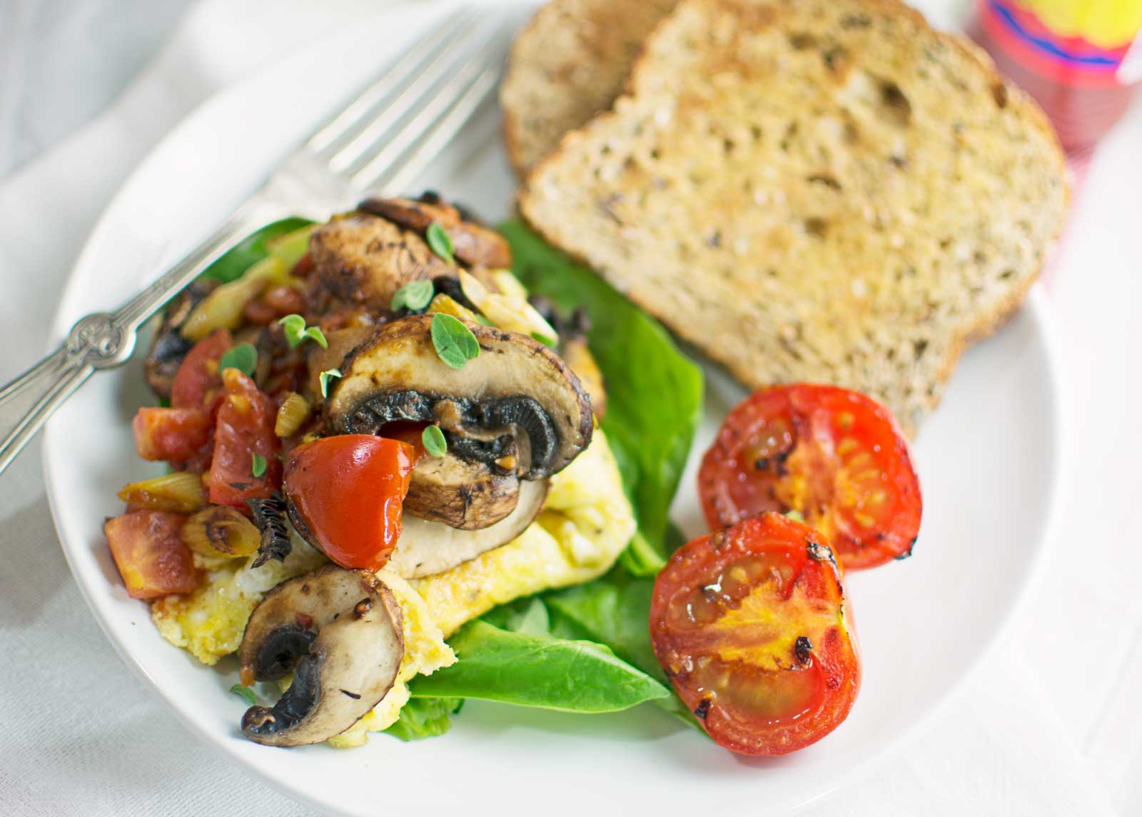 A plate with a fluffy omelette topped with mushrooms, cherry tomatoes, and herbs, served alongside fresh spinach, grilled tomato halves, and two slices of whole grain toast. A fork and a glass are partly visible in the background.