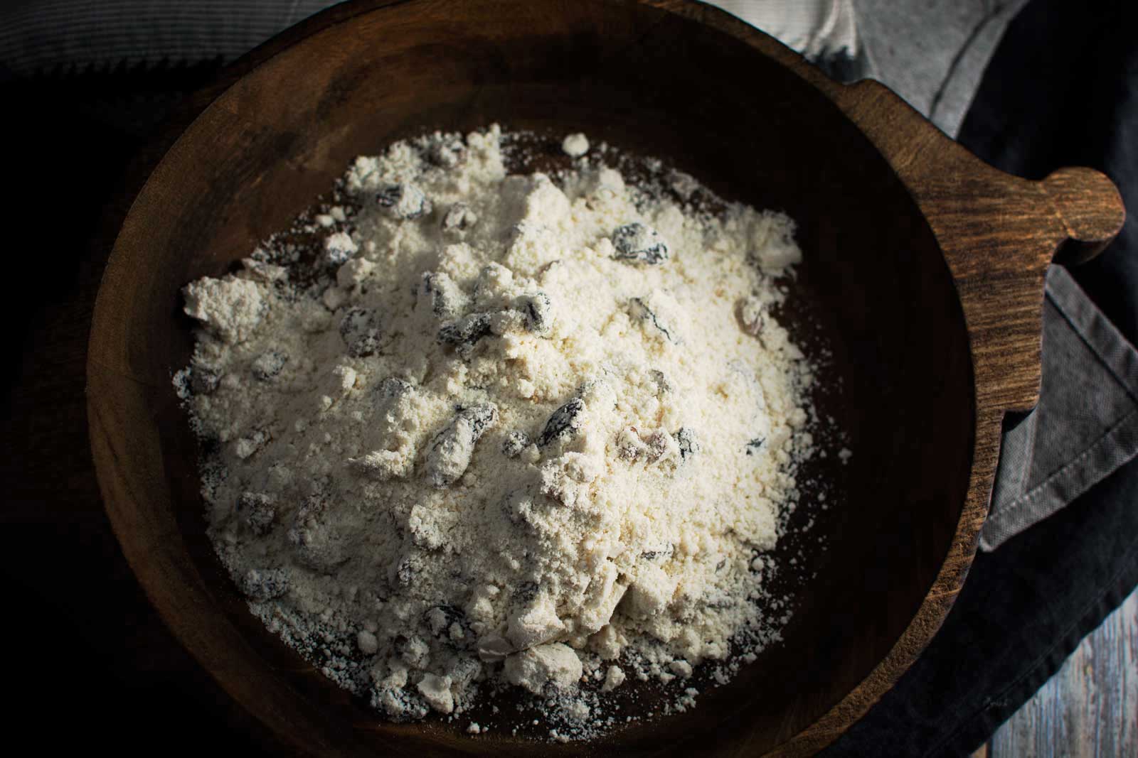 A wooden bowl filled with flour and small chunks, possibly raisins or nuts for raisin scone dough, sits on a dark surface with part of a gray cloth visible nearby.