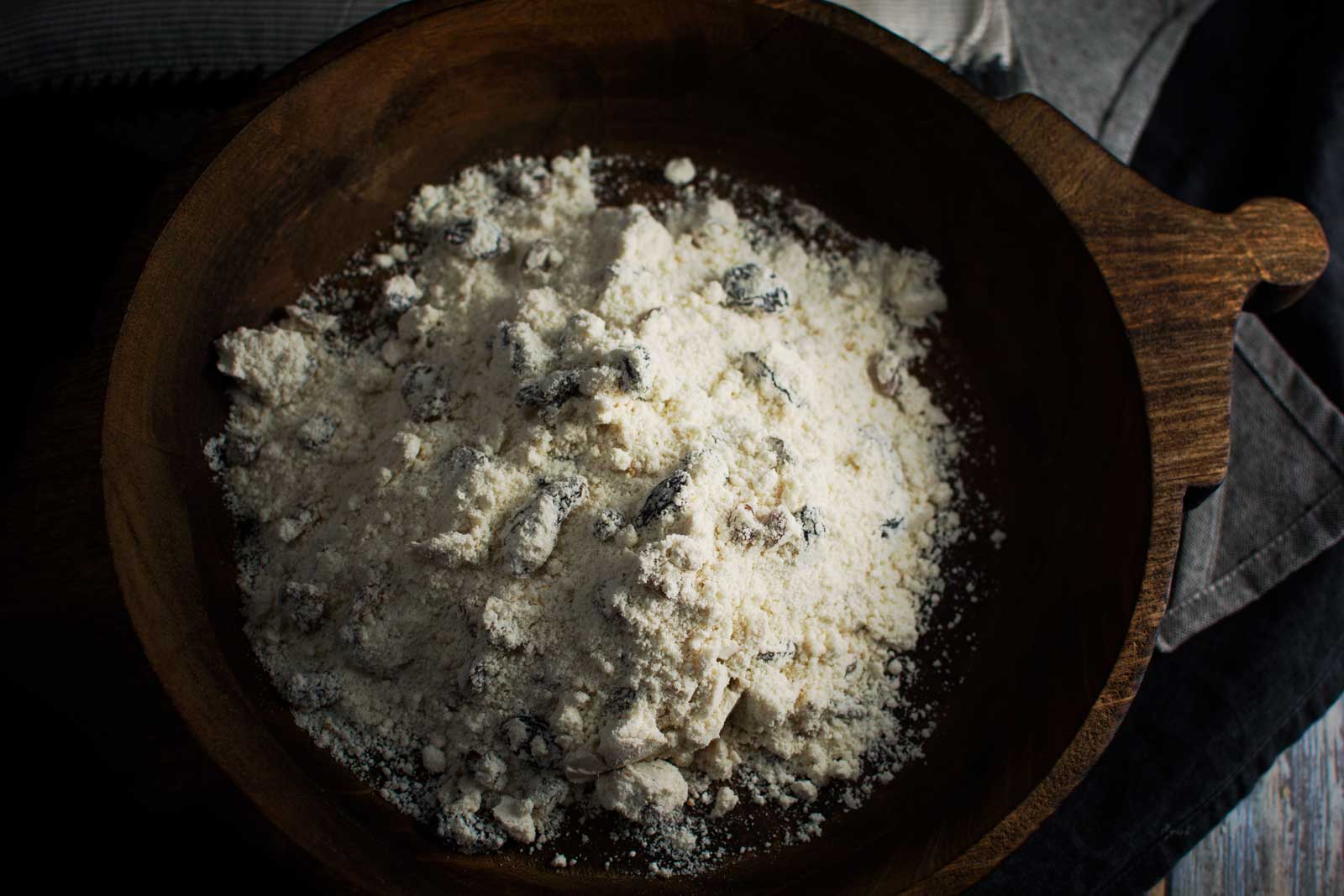 A wooden bowl filled with flour and raisins, partially mixed together for a delicious raisin scone, sits on a dark surface with a cloth nearby.