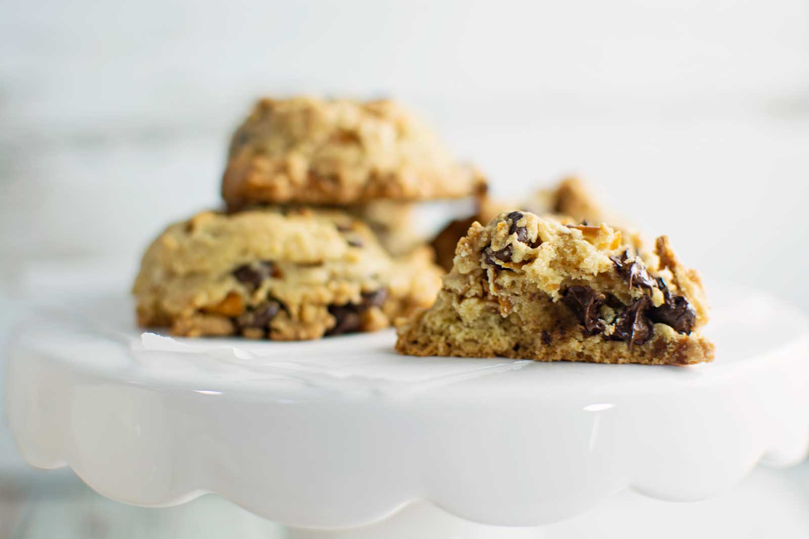 Three Chocolate Chip Pretzel Cookies rest on a white, scalloped cake stand. One cookie in the foreground is split open, revealing a soft, chewy texture with visible chocolate chips and bits of crunchy pretzel.