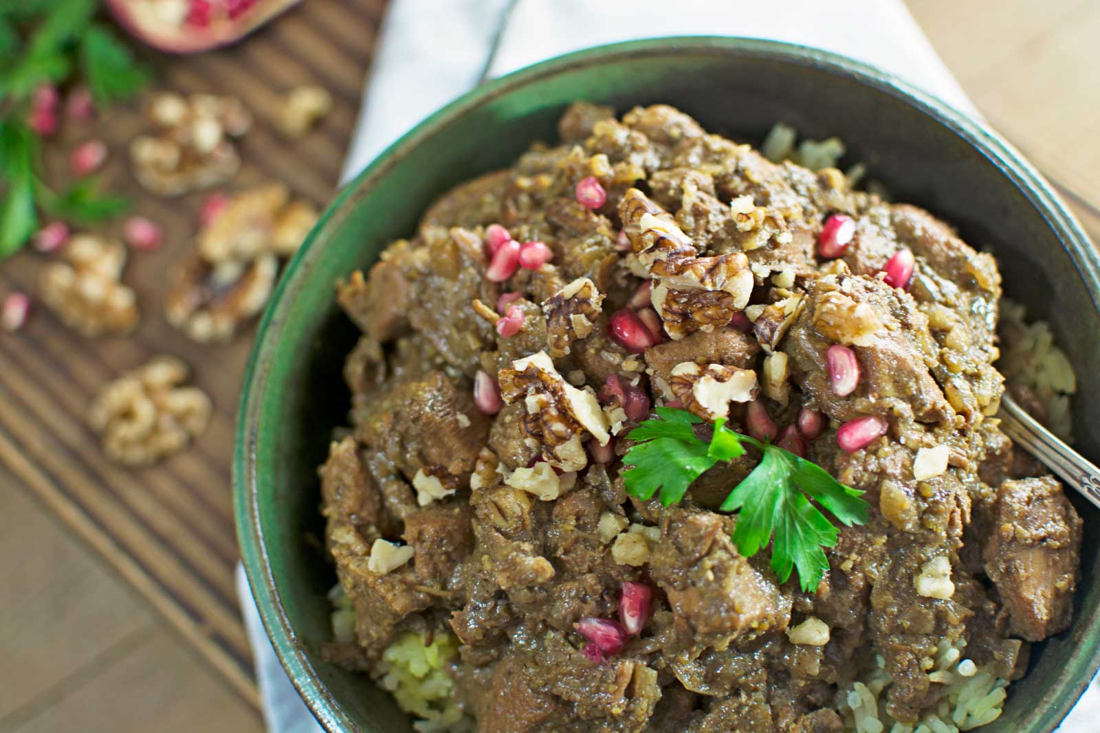 A bowl of Persian Fesenjan stew with pieces of meat in a rich brown sauce, garnished with chopped walnuts, pomegranate seeds, and a sprig of parsley, served over rice.