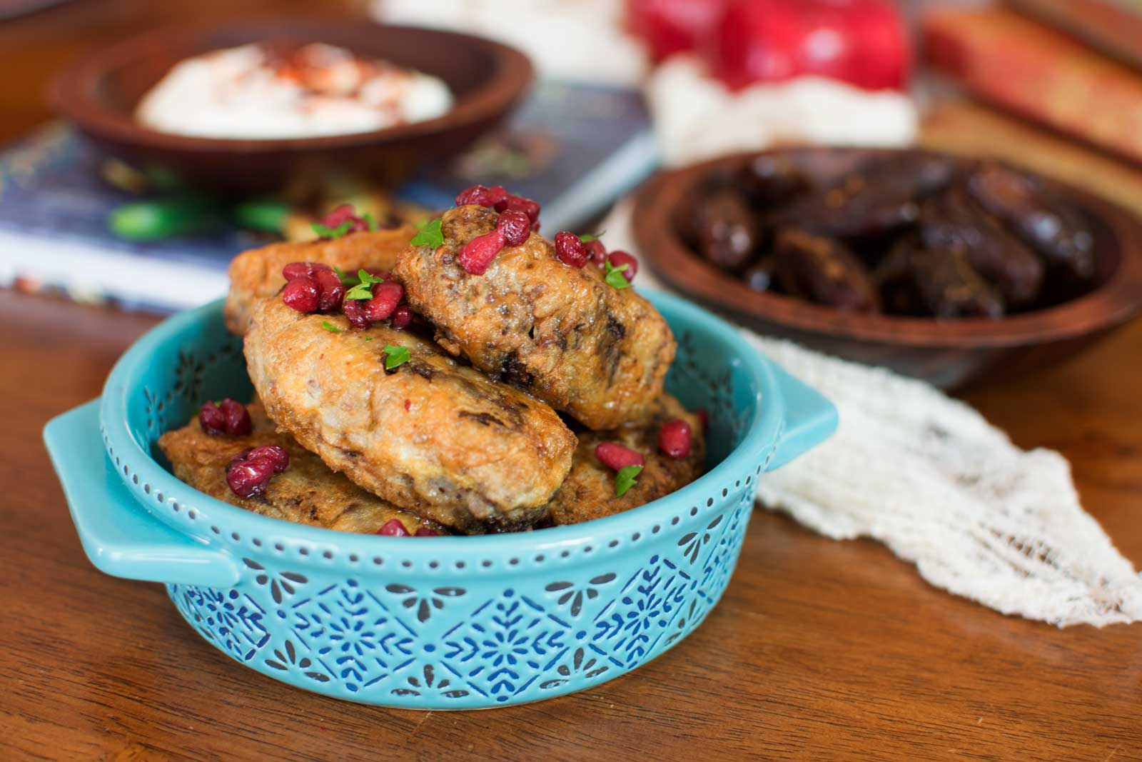 A blue bowl from the Bohemian Kitchen holds fried chicken wings garnished with pomegranate seeds and herbs. In the background, bowls of yogurt dip and dates rest on a wooden table.