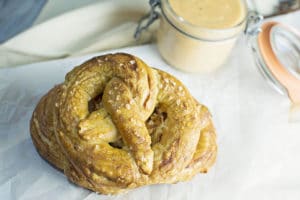 A golden-brown homemade pretzel sprinkled with coarse salt sits on parchment paper next to a jar of creamy dipping sauce.