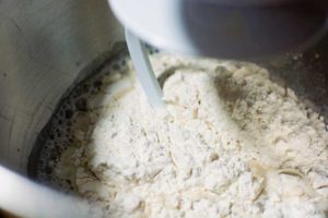 Close-up of flour, water, and yeast being mixed in a metal bowl with a dough hook attachment on a stand mixer, showing the initial stage of making homemade pretzels.