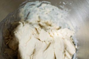 A close-up view of a ball of bread dough for homemade pretzels in a metal mixing bowl, with some flour visible on the sides of the bowl.
