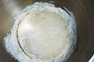 A ball of bread dough lightly dusted with flour sits in the center of a metal mixing bowl, ready to be shaped into homemade pretzels, with flour scattered around the edges.