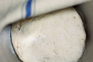 A ball of dough for homemade pretzels rises in a metal bowl, partially covered by a cloth with a blue stripe.