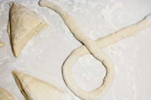 A homemade pretzel shaped into a loop with a twist lies on a floured surface next to several triangular dough pieces.
