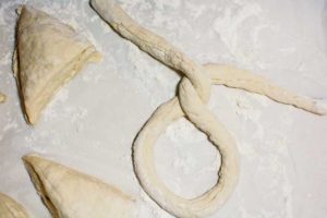 A piece of raw dough is being twisted into the shape of a Homemade Pretzel on a floured surface, with two triangular pieces of dough nearby.