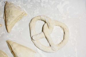 Homemade Pretzels dough is shaped into a classic pretzel, with a few triangular pieces nearby, all resting on a floured surface.