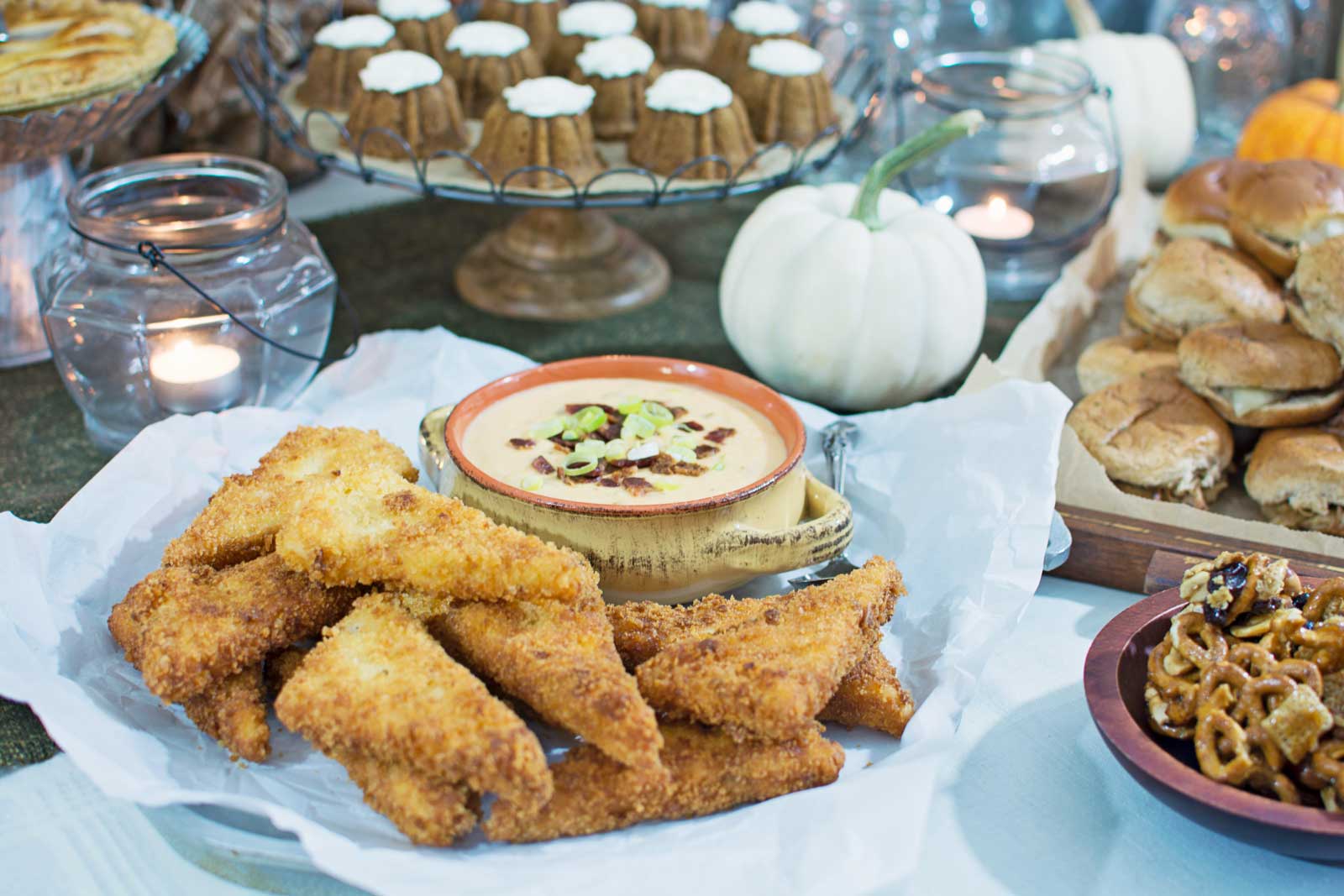 A platter of breaded fried fish filets with a bowl of dip, surrounded by mini cakes, dinner rolls, and pretzels sets the stage for a CMA Awards-inspired gathering, complete with candles and a white pumpkin on a beautifully decorated table.