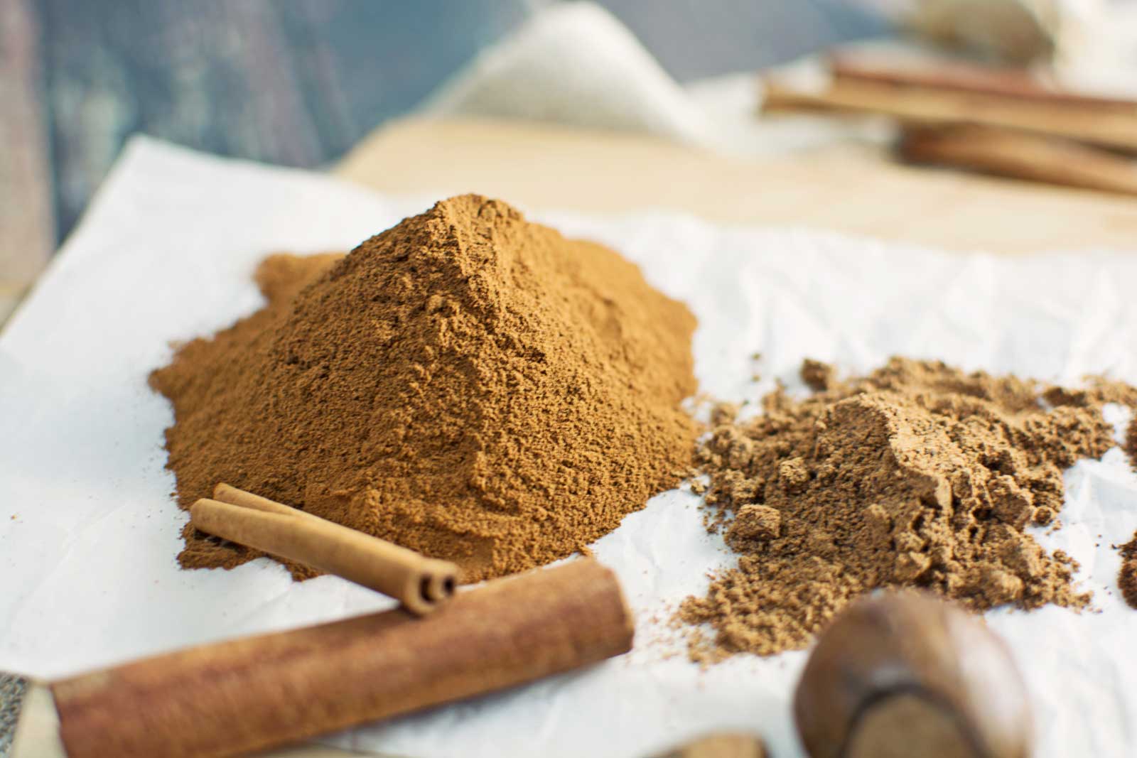 A close-up of two piles of ground brown spice, likely used for Pumpkin Pie, on white parchment paper. Whole cinnamon sticks and a whole nutmeg are placed nearby.