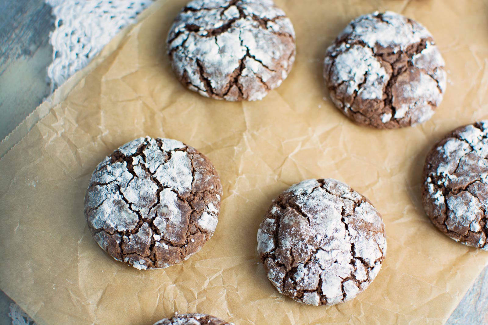 Six Crinkle Cookies dusted with powdered sugar rest on a sheet of parchment paper, their cracked surfaces revealing a soft, fudgy texture.