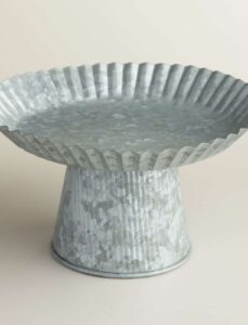 A galvanized metal cake stand with a fluted, scalloped edge and a sturdy, tapered base, displayed on a neutral background.