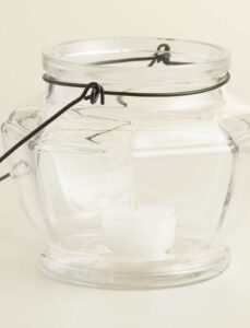 A clear glass lantern with a metal handle, containing a small white tealight candle inside, displayed against a plain light background.
