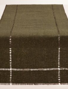 A dark olive green woven table runner with a subtle plaid pattern and openwork stitching, draped over the edge of a white table. The runner has short fringe along the ends.