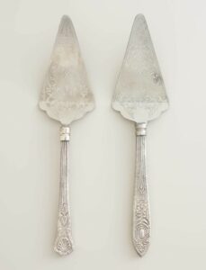 Two ornate silver pie servers with intricate engraved patterns on both the blades and the handles, placed side by side on a light-colored background.