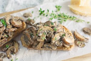 Two slices of toasted bread topped with creamy mushrooms, garnished with grated cheese and fresh herbs, are placed on parchment paper with extra mushrooms and cheese in the background.