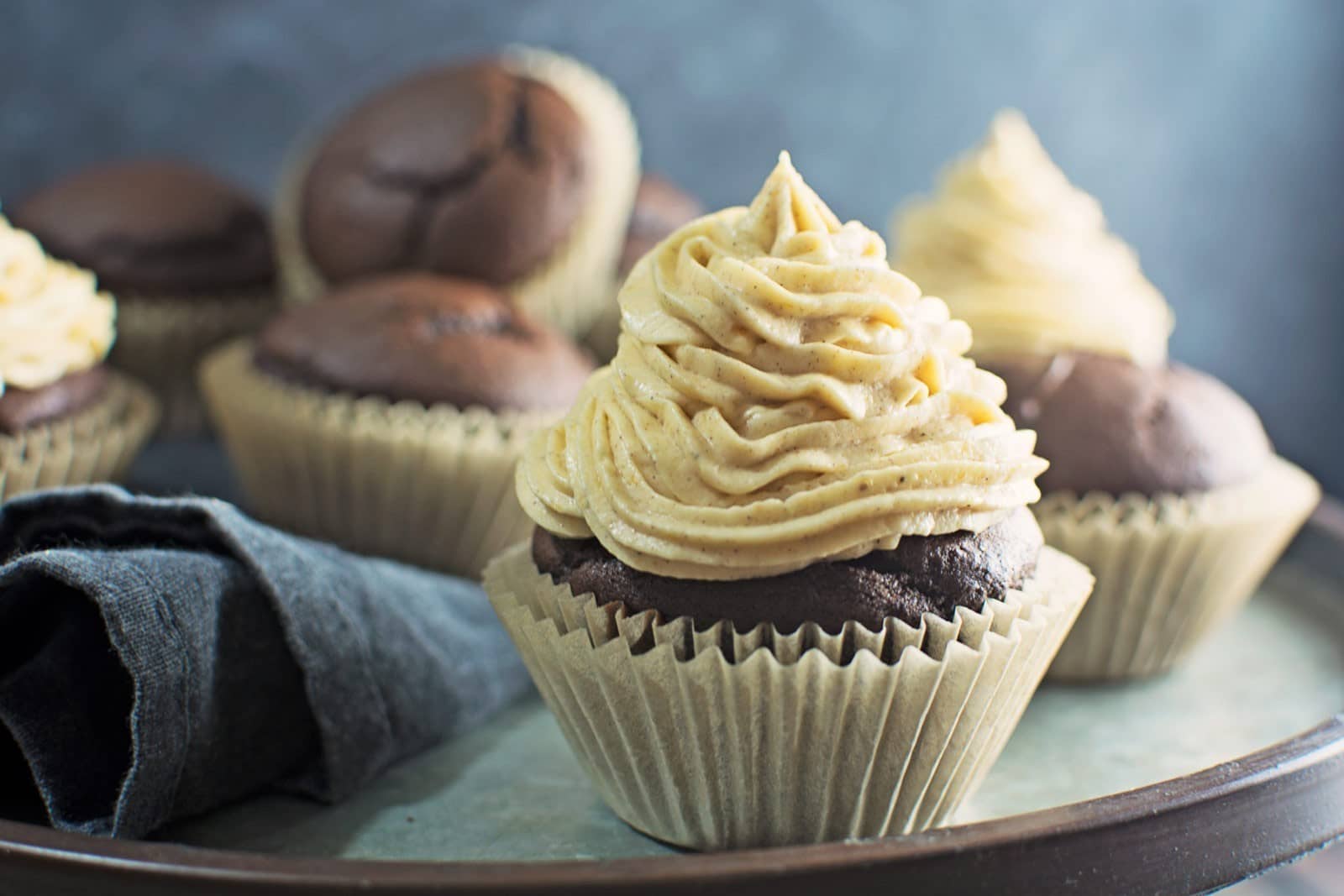 A close-up of rich chocolate cupcakes with creamy, swirled frosting on top, arranged on a tray with a dark napkin beside them against a blurred background. Perfect for cupcake lovers.