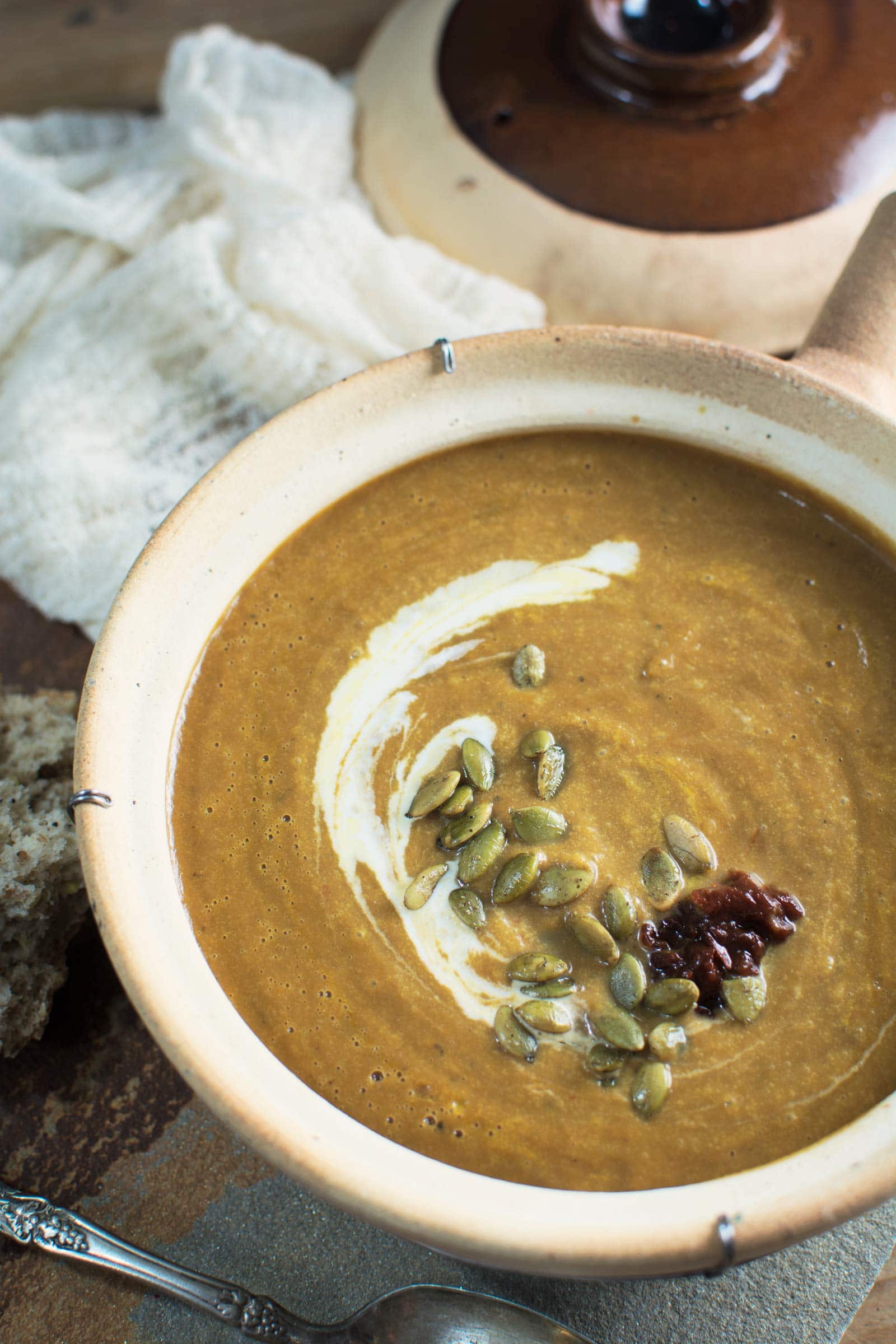 A bowl of creamy Pumpkin Soup garnished with a swirl of cream, pumpkin seeds, and a dollop of dark paste sits beside rustic bread and a cloth on a wooden surface.