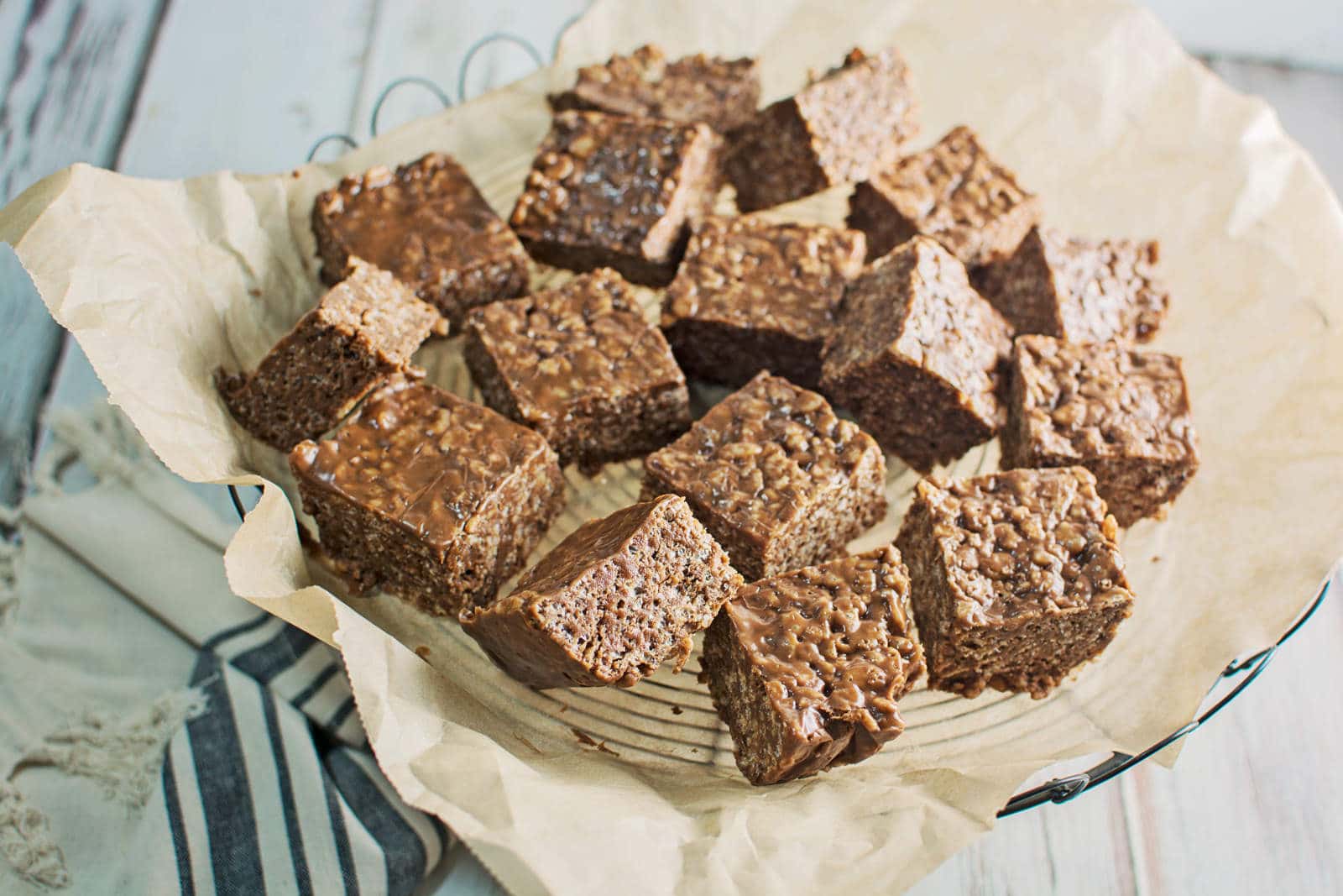 A basket lined with parchment paper holds several chocolate caramel rice crispy squares, neatly cut into bite-sized pieces. The treats have a textured, crunchy appearance and are arranged in a casual pile.