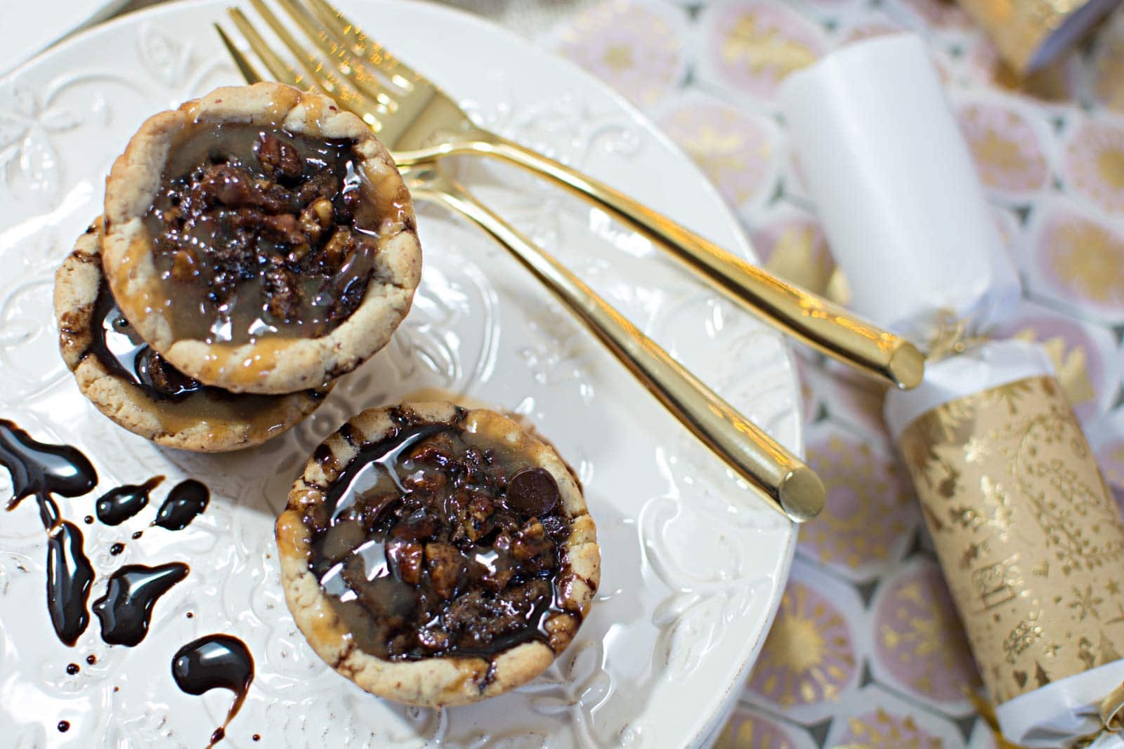 Two small chocolate nut tassies sit on a white plate, drizzled with dark sauce. Gold utensils and a festive gold and white party cracker are placed beside the plate on a patterned surface.