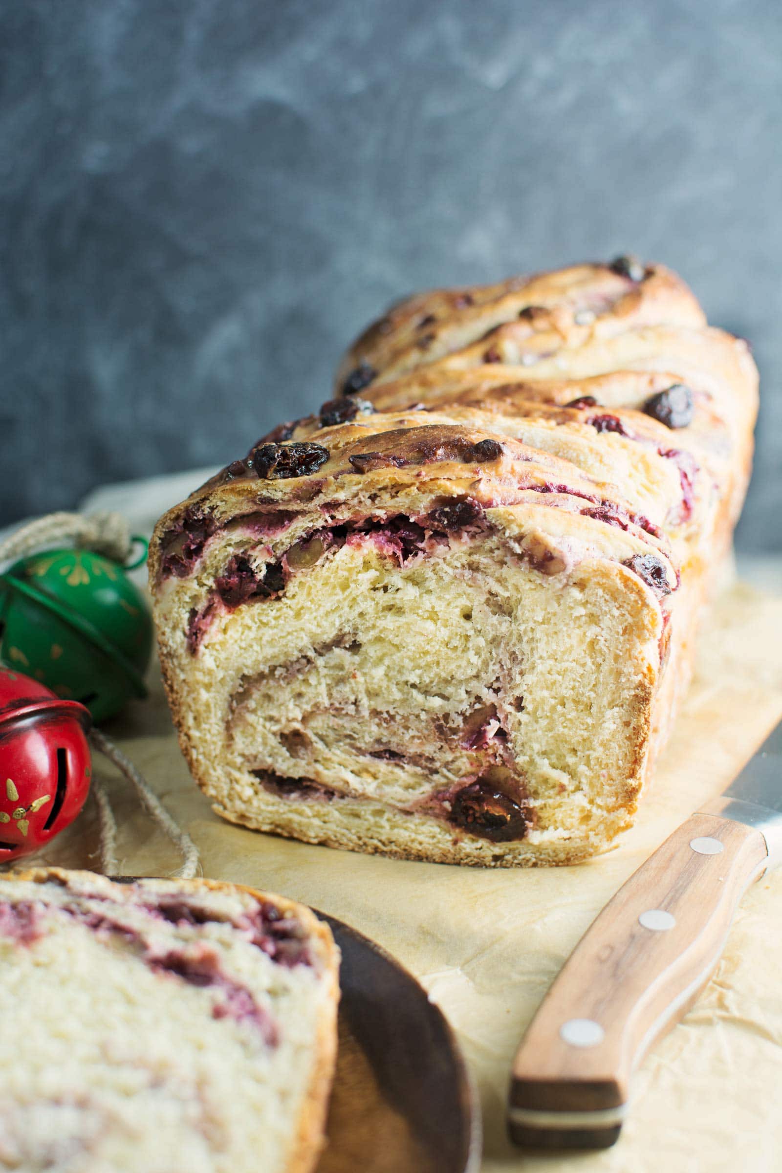 A loaf of swirled babka with visible berries and raisins is partially sliced on a cutting board next to a wooden-handled knife. Two holiday jingle bells are in the background.