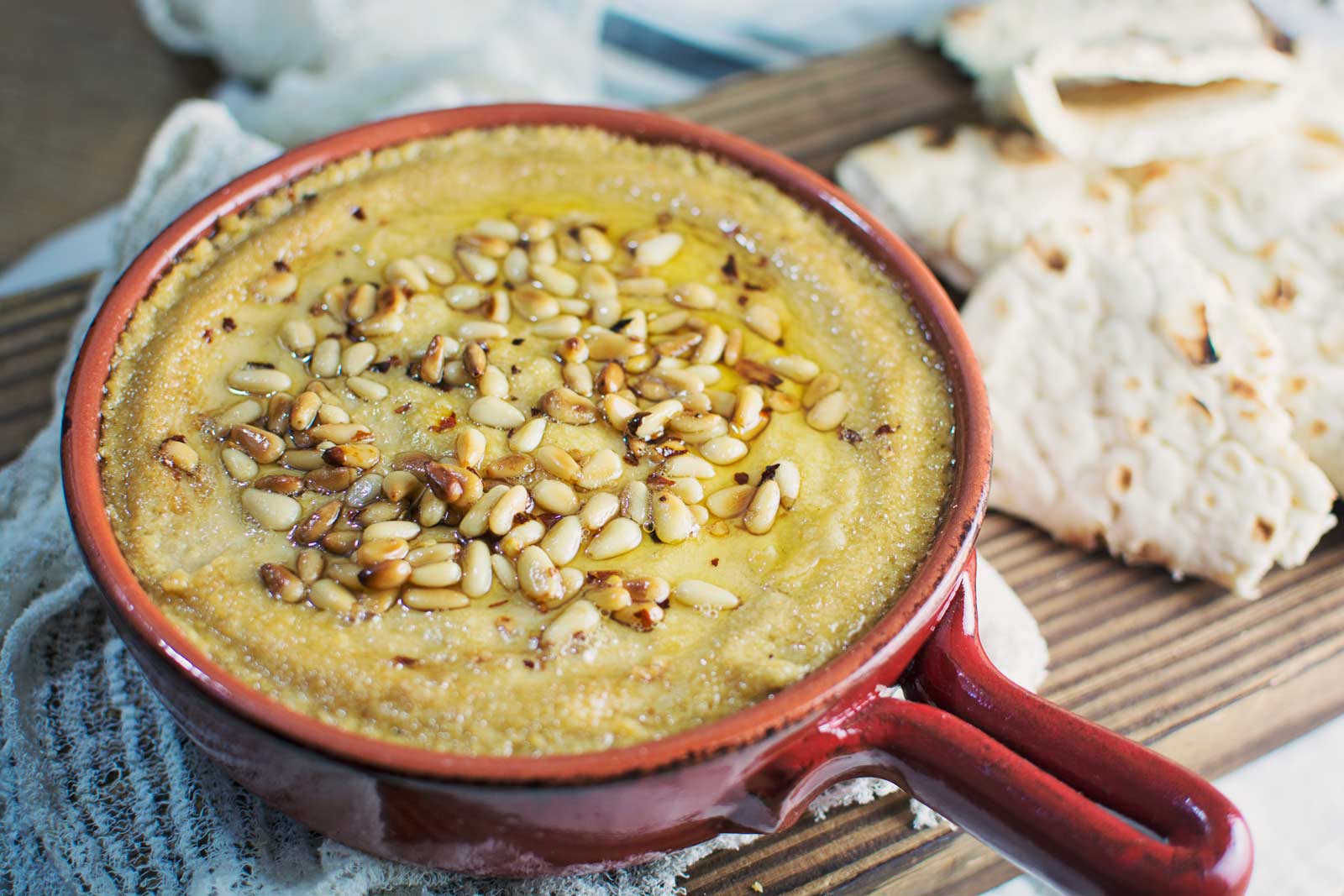 A red dish filled with creamy baked hummus topped with pine nuts and olive oil, placed on a textured cloth next to pieces of flatbread on a wooden board.