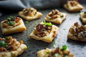 Small, square shallot tarts topped with sautéed mushrooms and fresh parsley are arranged on a dark, textured tray.