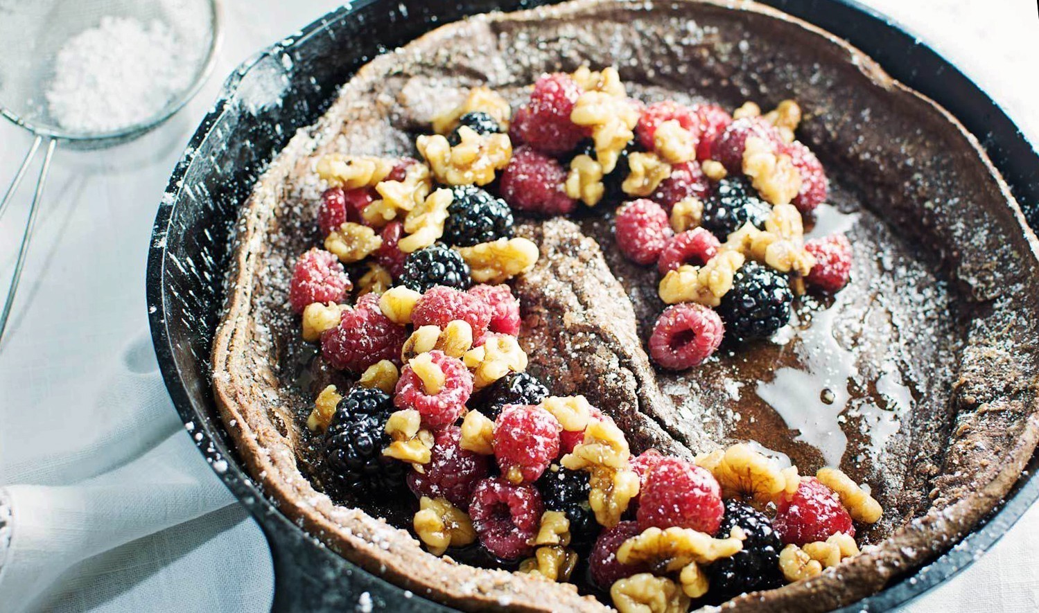 A Chocolate Dutch Baby pancake in a skillet, topped with fresh raspberries, blackberries, walnuts, and a dusting of powdered sugar. Nearby, a metal sieve holds extra sweetness for this indulgent Chocolate Dutch Baby treat.