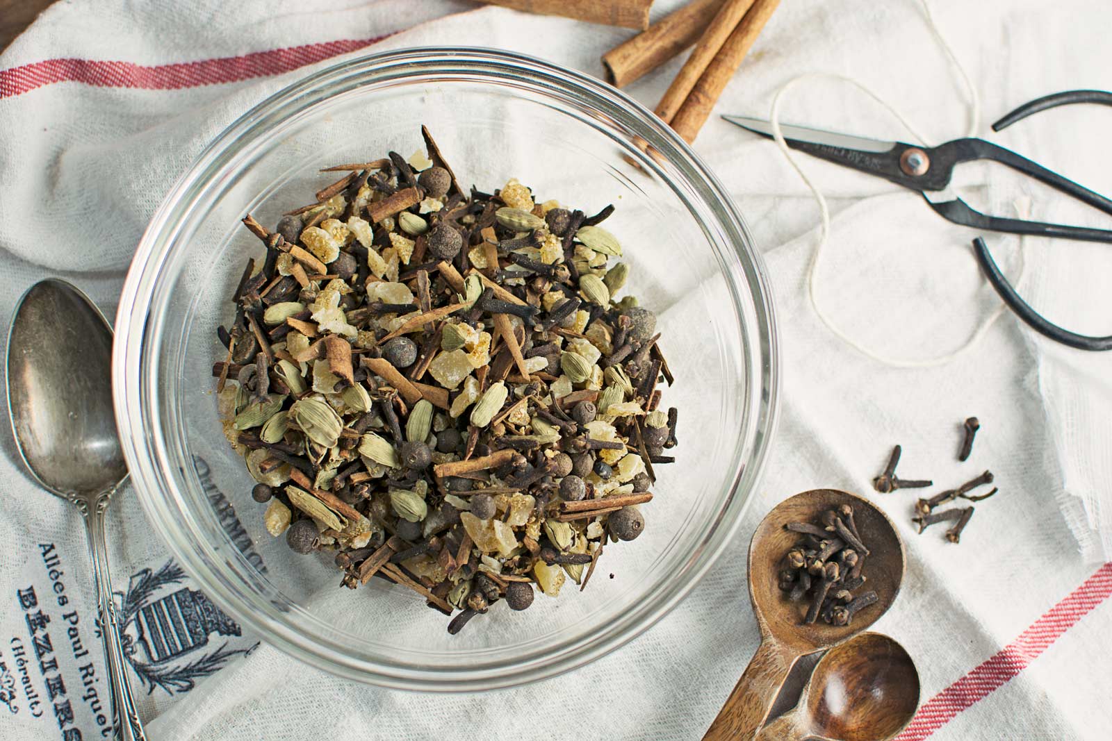 A glass bowl filled with mixed whole mulling spices, including cardamom, cloves, allspice, and cinnamon sticks, sits on a white cloth next to a spoon, wooden spoon, scattered cloves, scissors, and string.