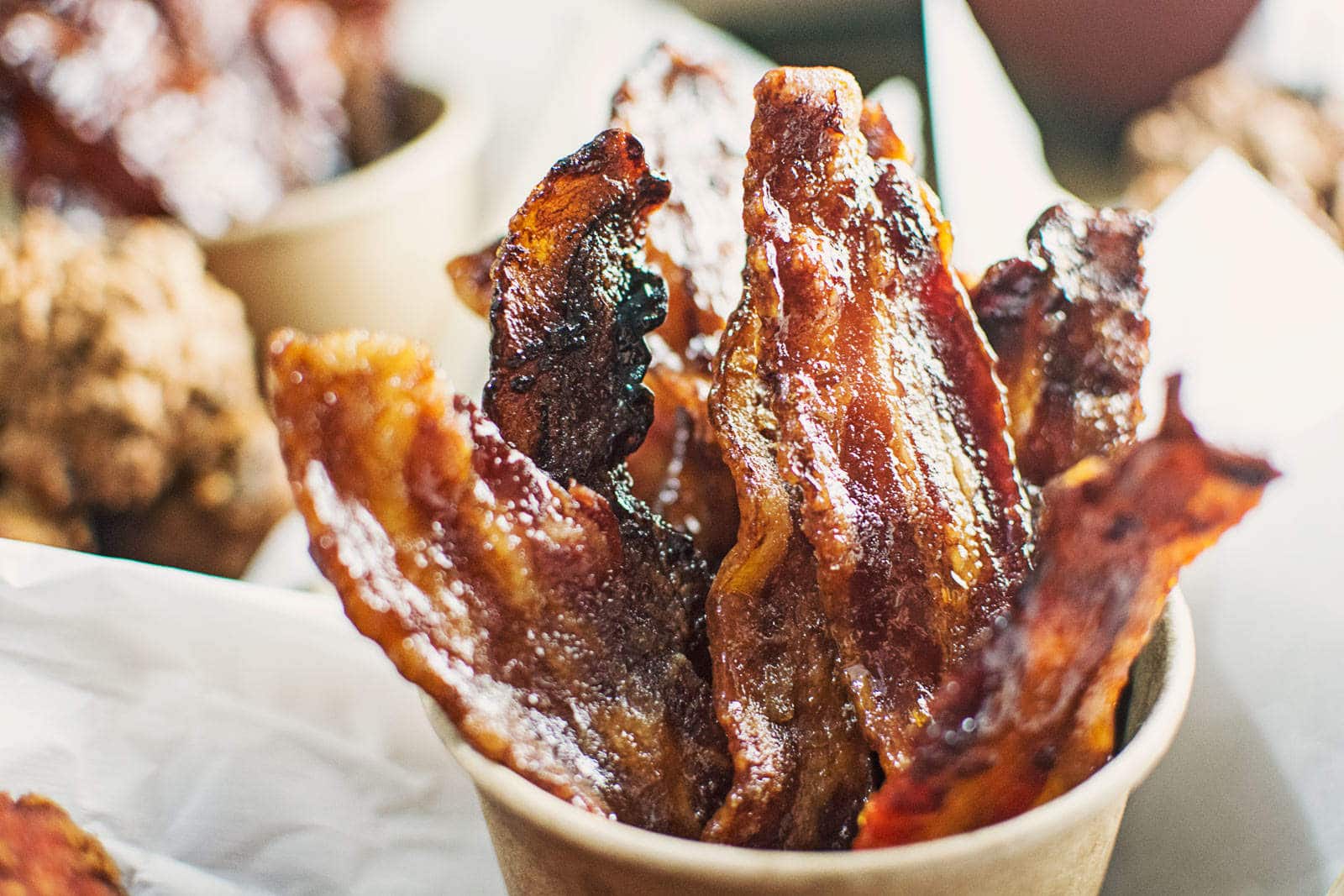 Crispy, caramelized strips of Pig Candy bacon are arranged upright in a brown paper cup, glistening with a shiny glaze. The background shows blurred food items.