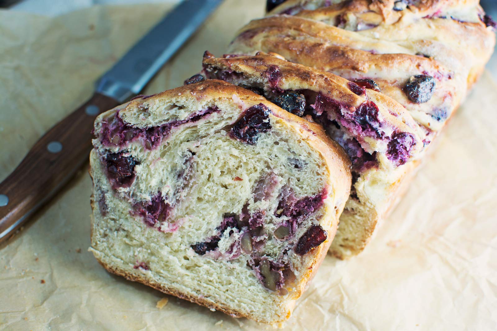 A loaf of braided babka with visible swirls of berry filling and nuts, sliced to reveal the moist interior, sits on parchment paper next to a wooden-handled bread knife.