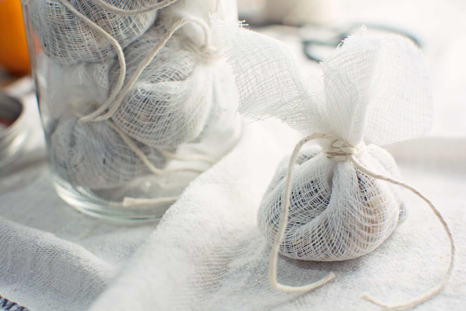 A small bundle of mulling spices wrapped in white cheesecloth and tied with string sits on a white cloth, with similar bundles visible inside a glass jar in the background.