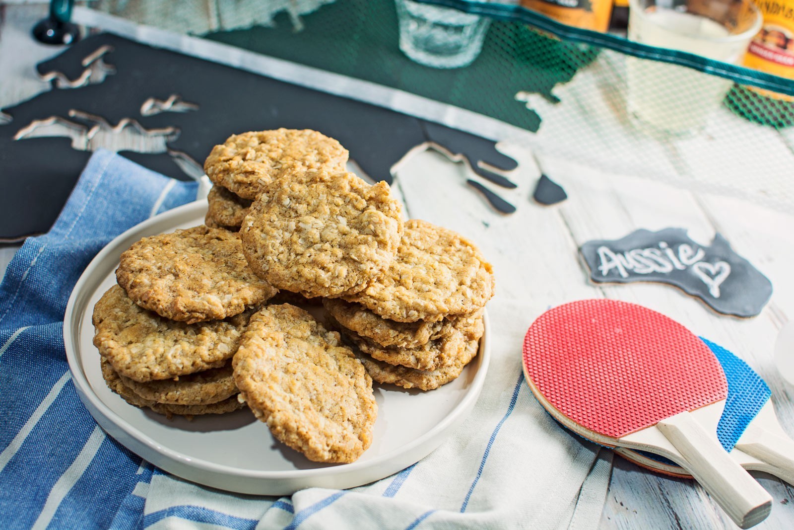 A plate of Anzacs oatmeal cookies is stacked on a striped cloth next to two ping pong paddles and a small chalkboard sign that says Aussie with a heart. A metal map of Australia is in the background.