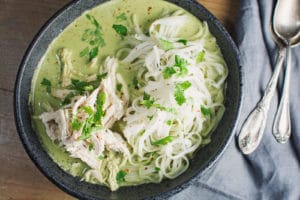 A bowl of Thai Chicken green curry soup with shredded chicken, rice noodles, and fresh herbs, served on a wooden table next to a gray napkin and two metal spoons.