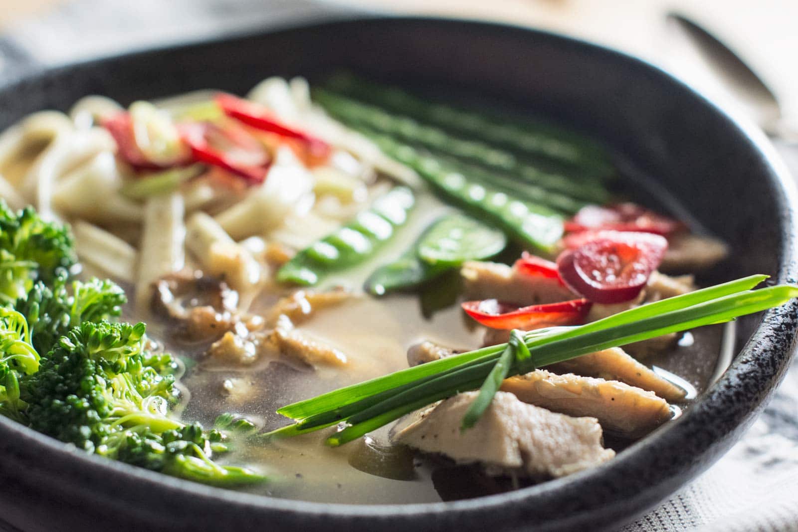 A bowl of Noodle Soup with sliced chicken, broccoli, snow peas, cherry tomatoes, red chili slices, and green onions in a clear broth, served in a black bowl.