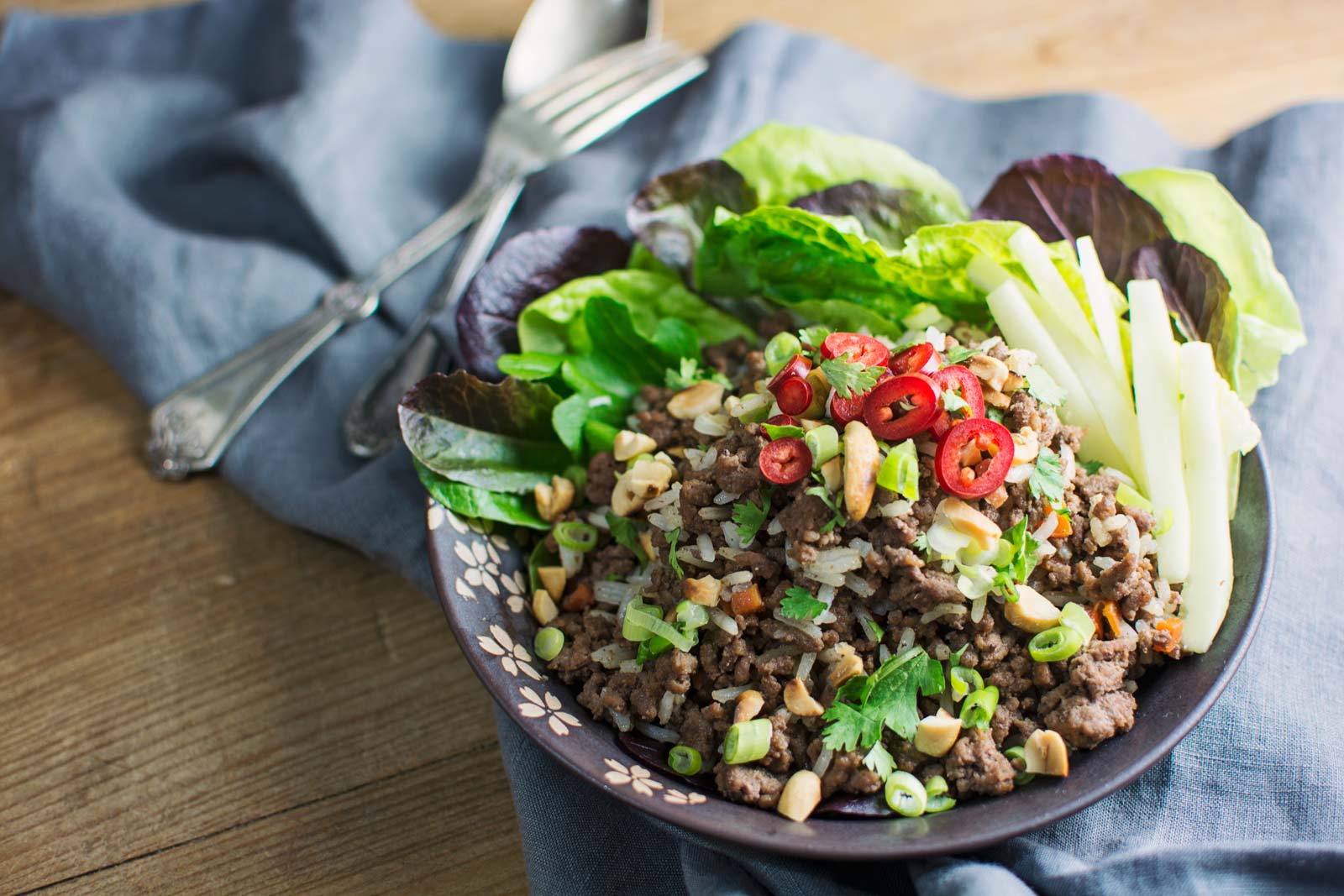 A bowl of Ginger Beef topped with chopped green onions, red chili slices, peanuts, and herbs, served with lettuce leaves and cucumber sticks, placed on a wooden table with a gray cloth and silverware in the background.