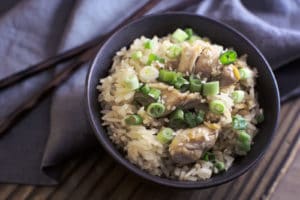 A bowl of Chicken Rice topped with chopped green onions and sesame seeds sits on a wooden table, with chopsticks and a gray napkin beside it.