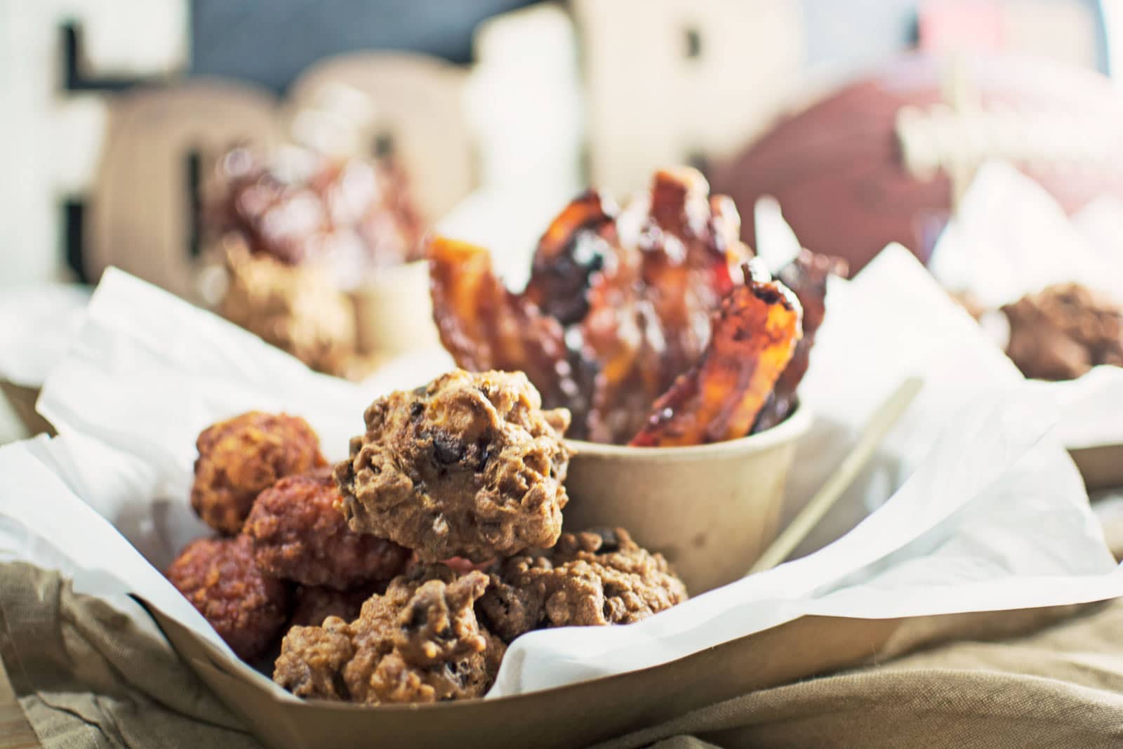 A tray lined with parchment paper holds spicy chicken wings, crispy bacon strips in a bowl, Hot Dog Fritters, and clusters of cookies, set on a table with a football and blurred decorations in the background.