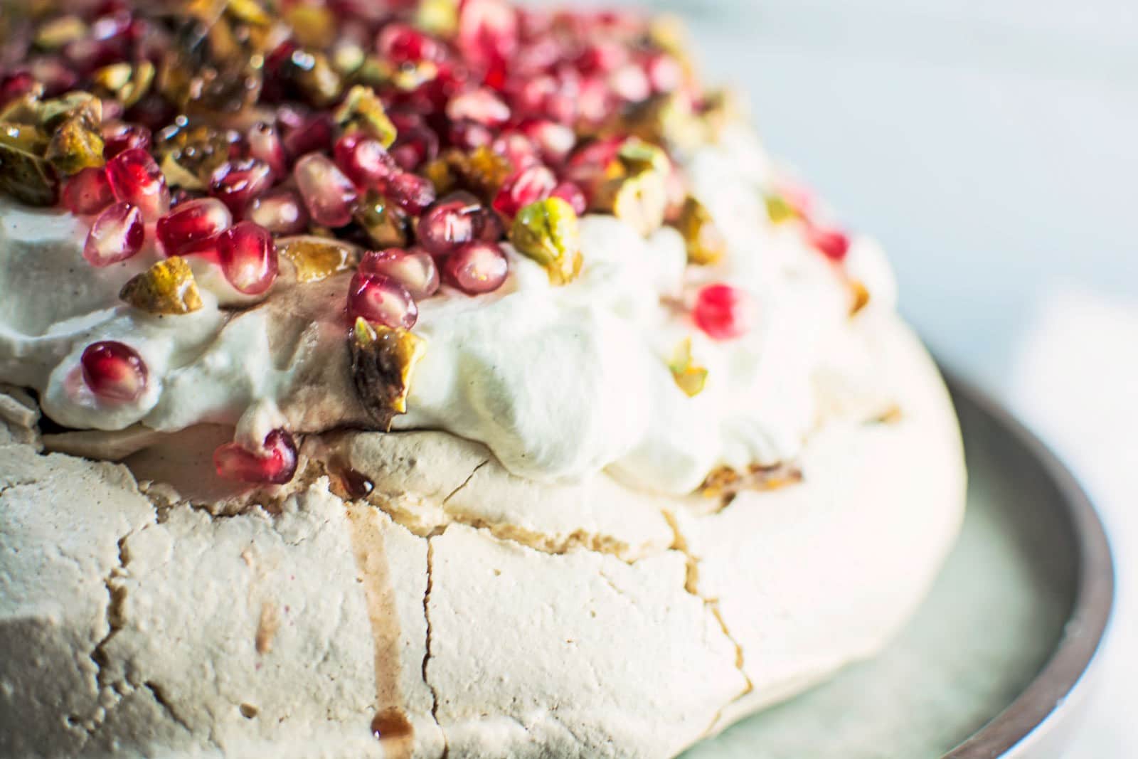 Close-up of a Winter Pavlova dessert topped with whipped cream, pomegranate seeds, chopped pistachios, and a drizzle of syrup, sitting on a metal tray.